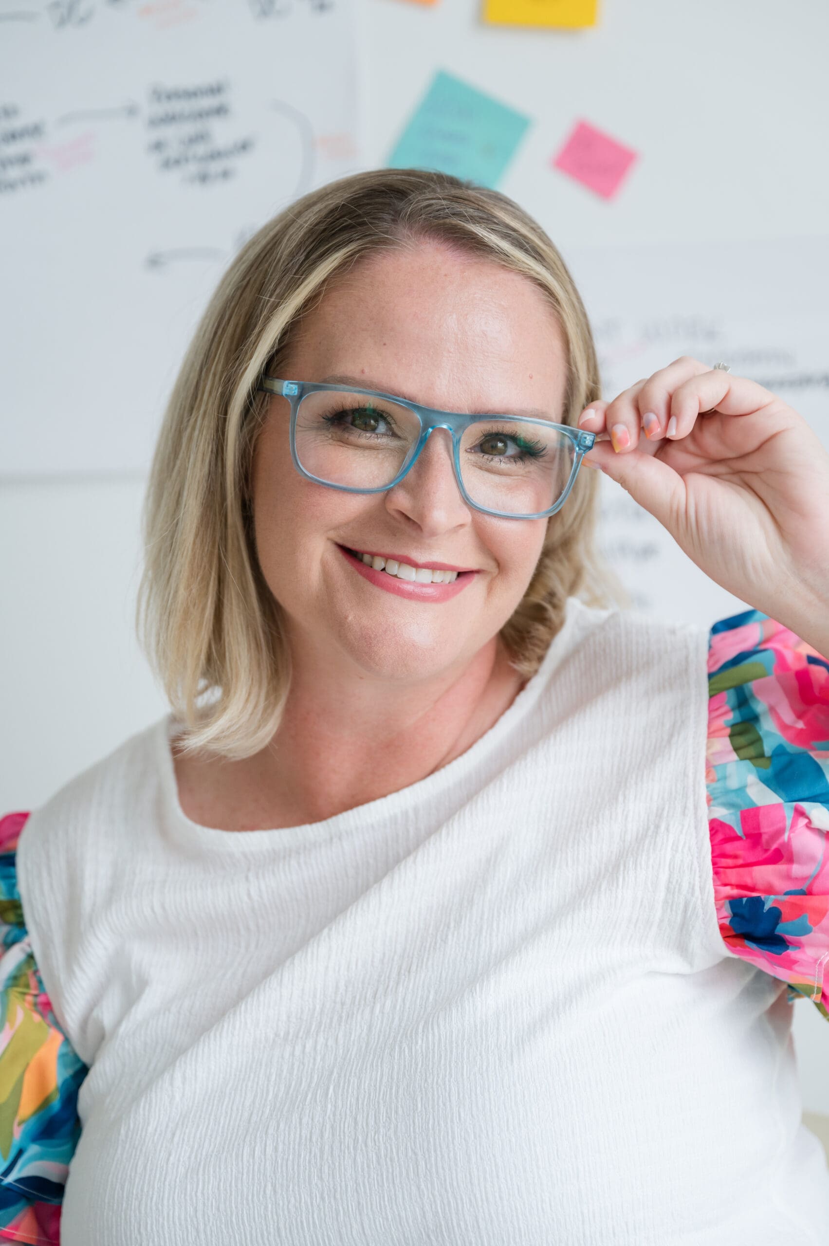 Smiling woman wearing blue glasses and a white top with colorful floral sleeves, posing confidently in front of a wall covered in sticky notes and workflow charts—representing creative business strategy and systems planning.