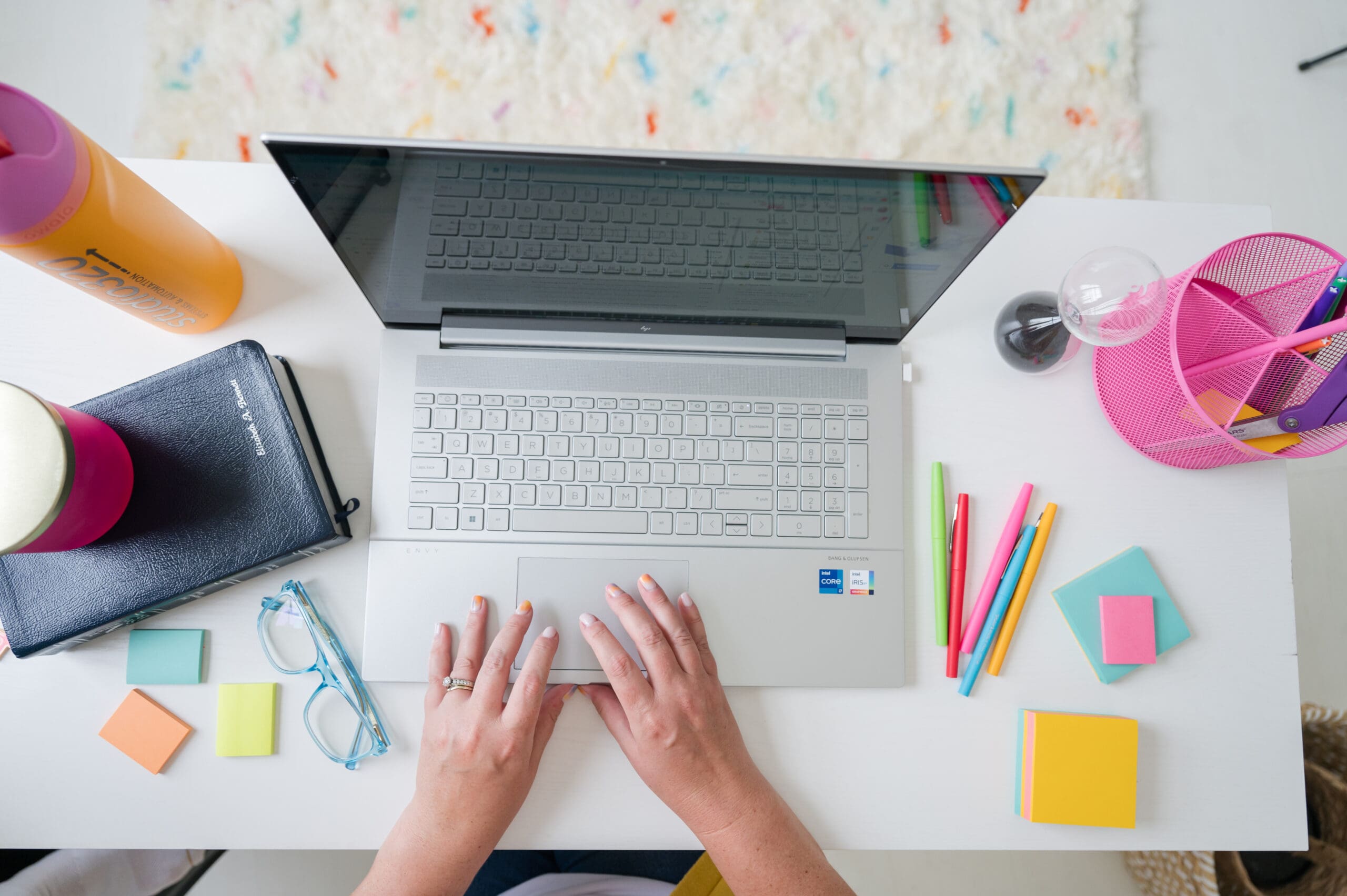 Top-down view of a woman working on a silver laptop at a bright, colorful desk with sticky notes, pens, glasses, a Bible, and a Studio320 water bottle—capturing a creative and faith-driven workspace for business planning and automation.