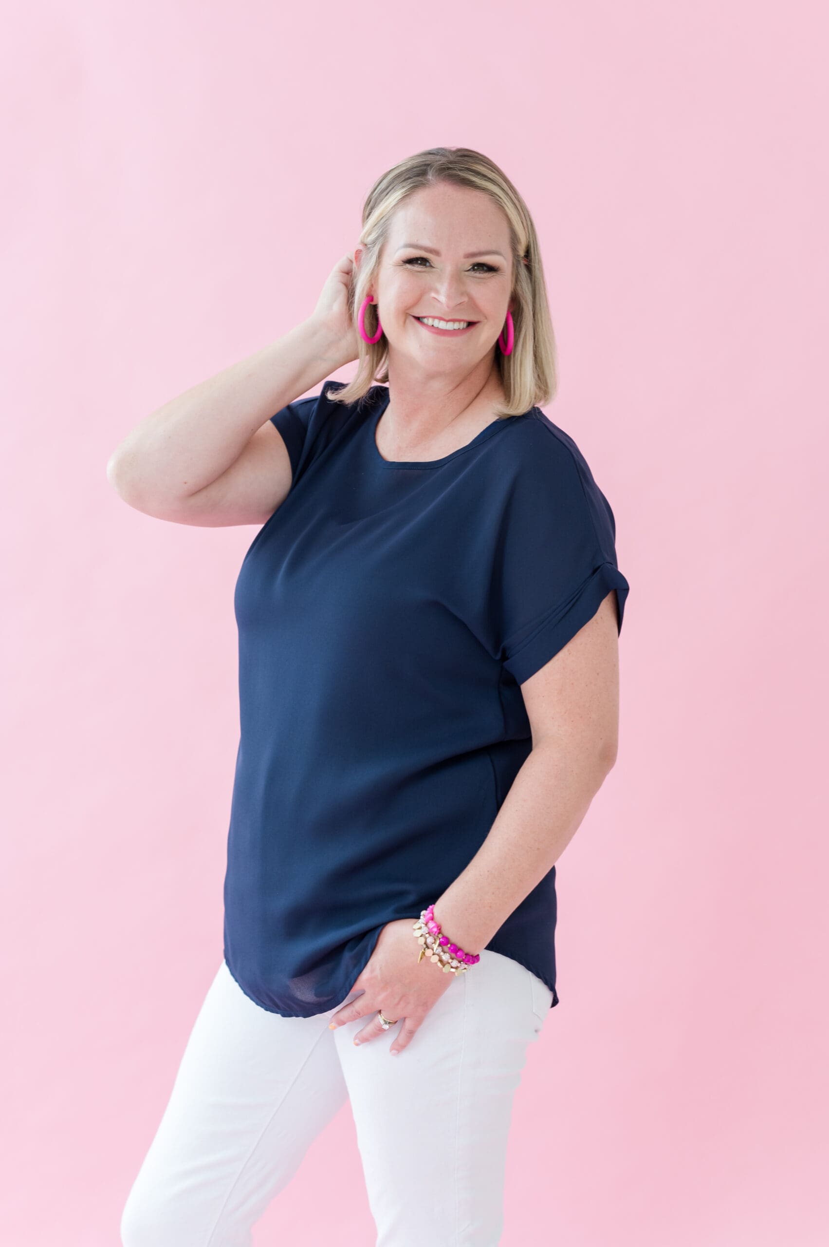 Smiling woman in a navy blouse and white pants posing confidently against a pink background, accessorized with bright pink earrings and bracelets—professional branding photo for a creative entrepreneur.