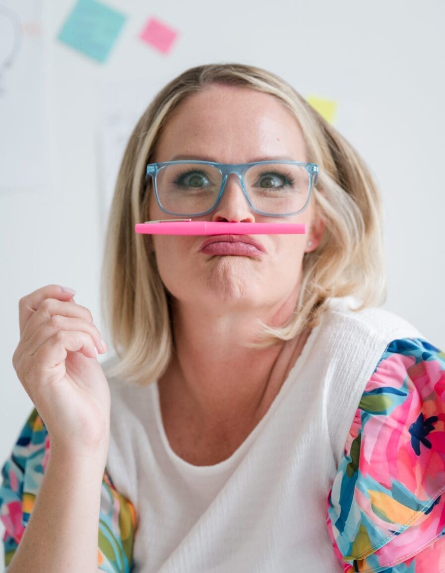 Playful woman in blue glasses making a funny face with a pink pen under her nose, sitting in a bright workspace with colorful sticky notes behind her.
