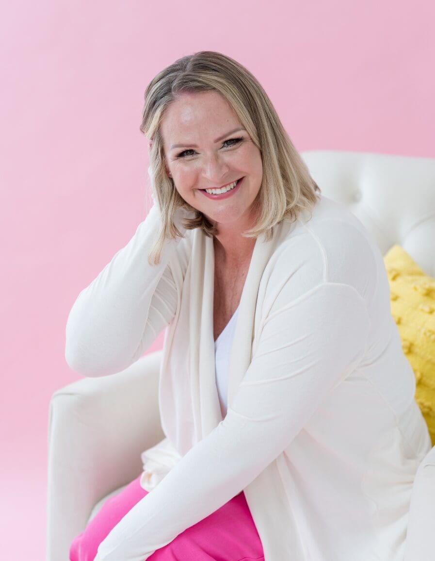 Friendly woman smiling while seated in a white chair, wearing a white cardigan and bright pink pants against a soft pink background.