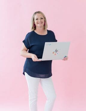 Friendly woman standing with an open laptop against a soft pink background, wearing a navy blouse, white pants, and bright pink earrings.