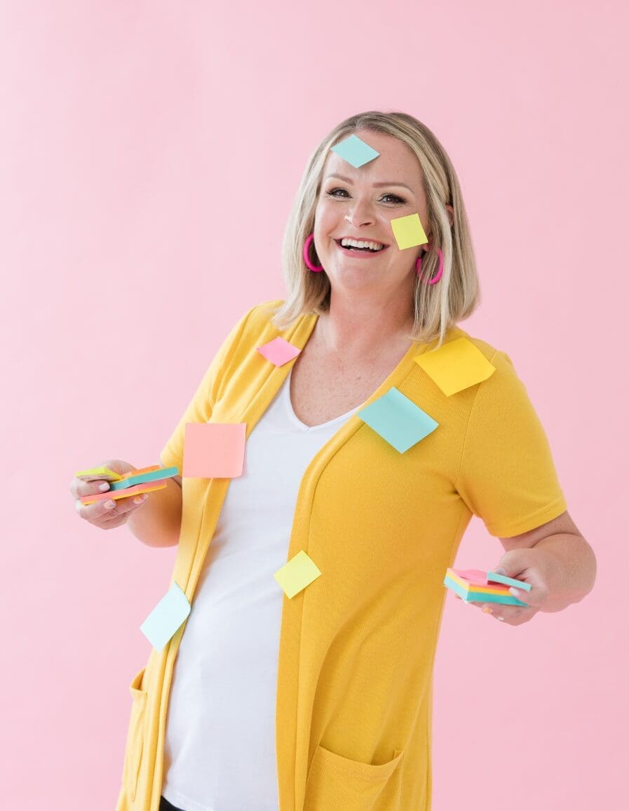 Cheerful woman in a yellow cardigan holding colorful sticky notes, with more notes playfully stuck to her outfit and face, against a bright pink background.